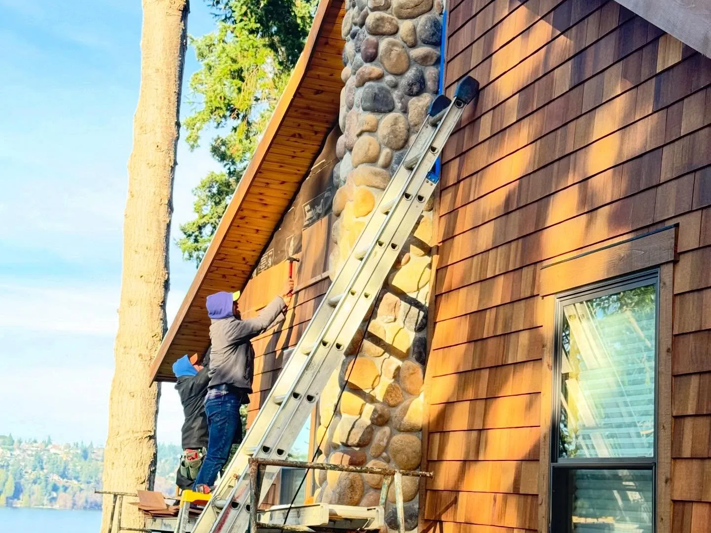 Crew installing cedar shingle siding beside a stone chimney, with the water behind