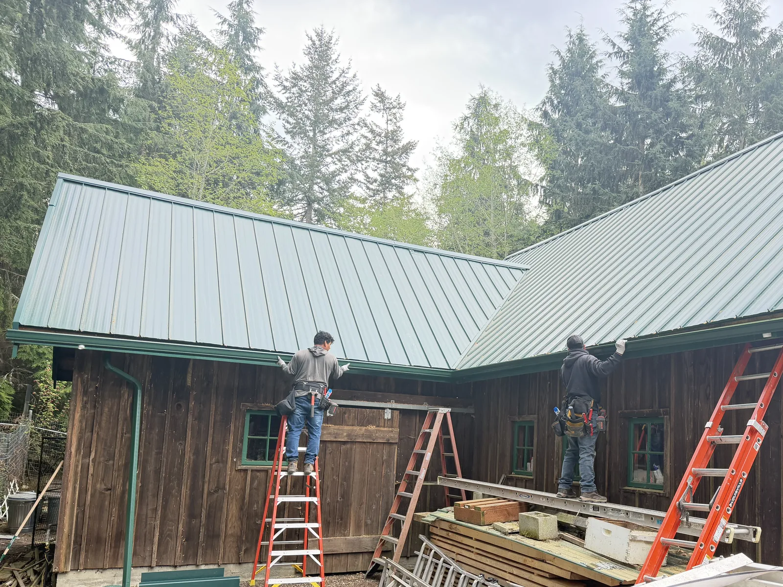 Two installers on ladders hanging green gutters under a new standing-seam metal roof