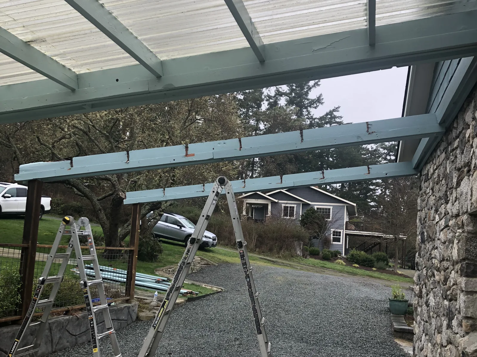 Patio cover frame with roof panels going on and ladders on the gravel below