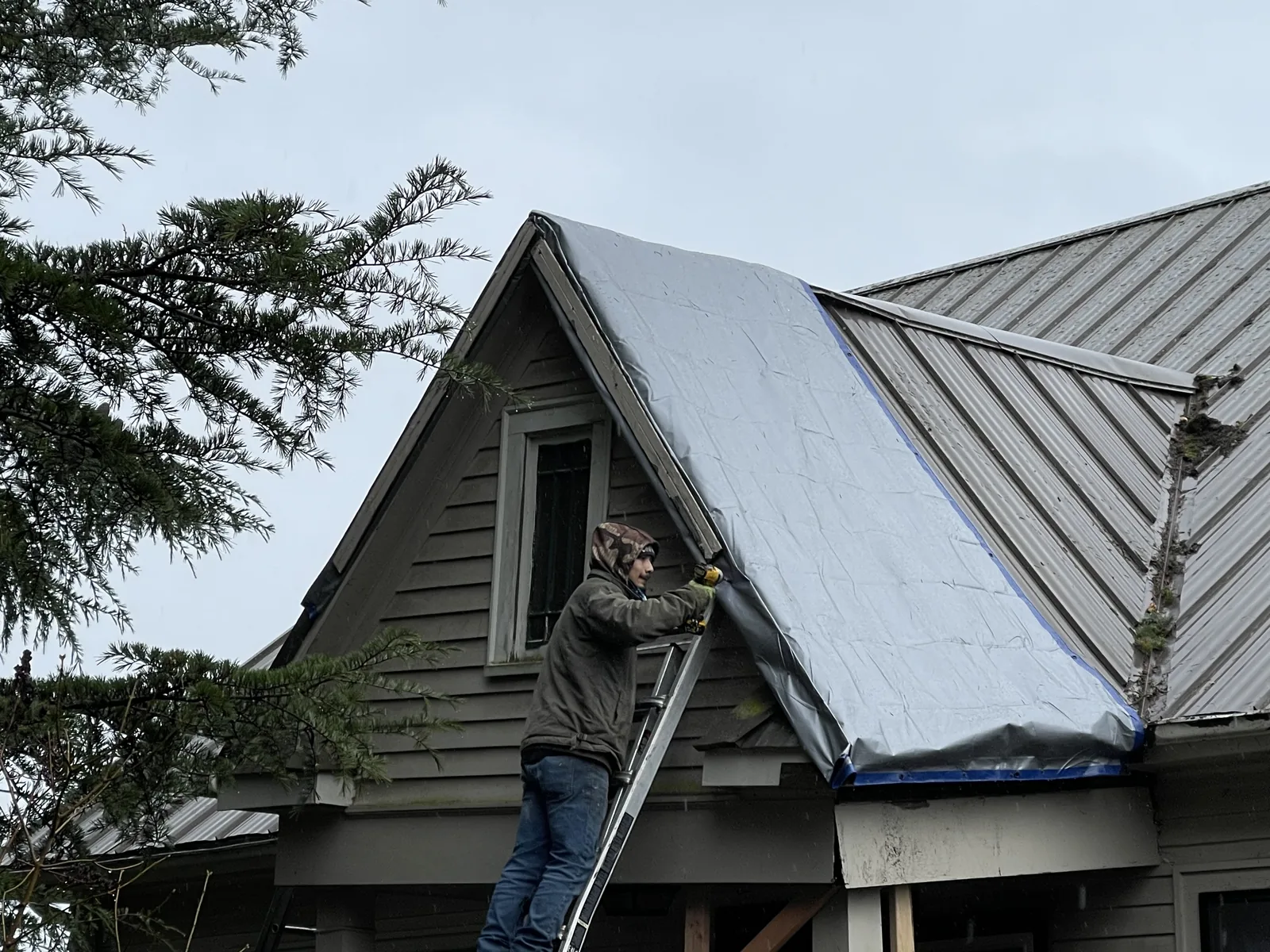 Roofer on a ladder covering a storm-damaged gable with a tarp