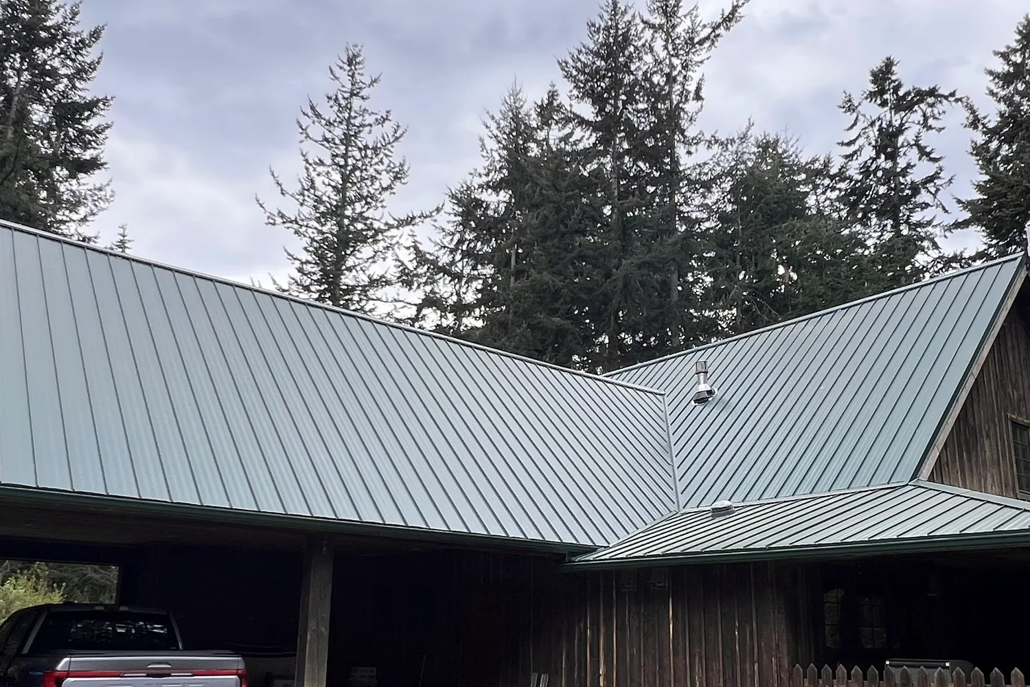 New grey-green standing-seam metal roof over a home and attached carport, fir trees behind