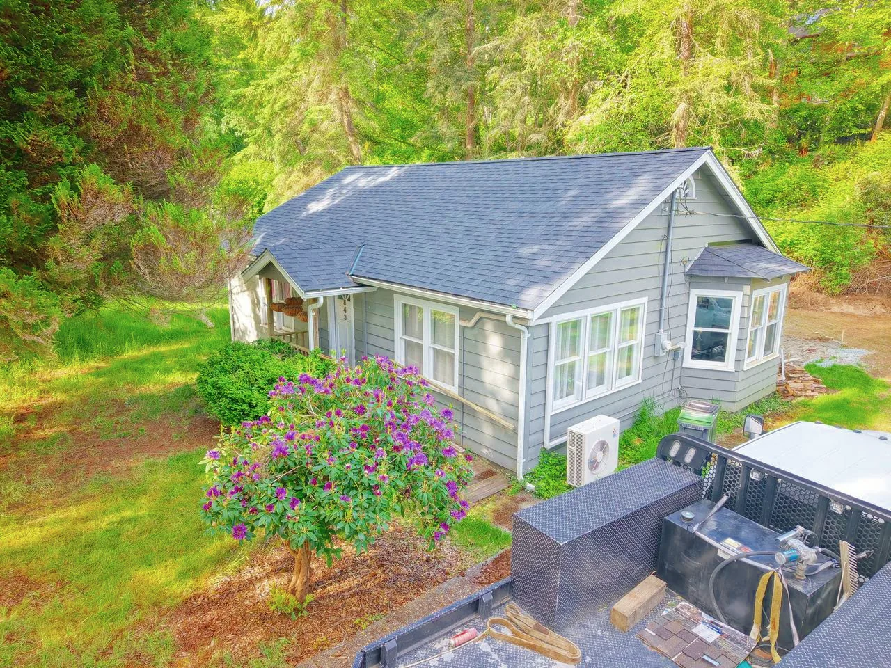 Drone view of a small grey cottage with a shingle roof and a flowering shrub out front