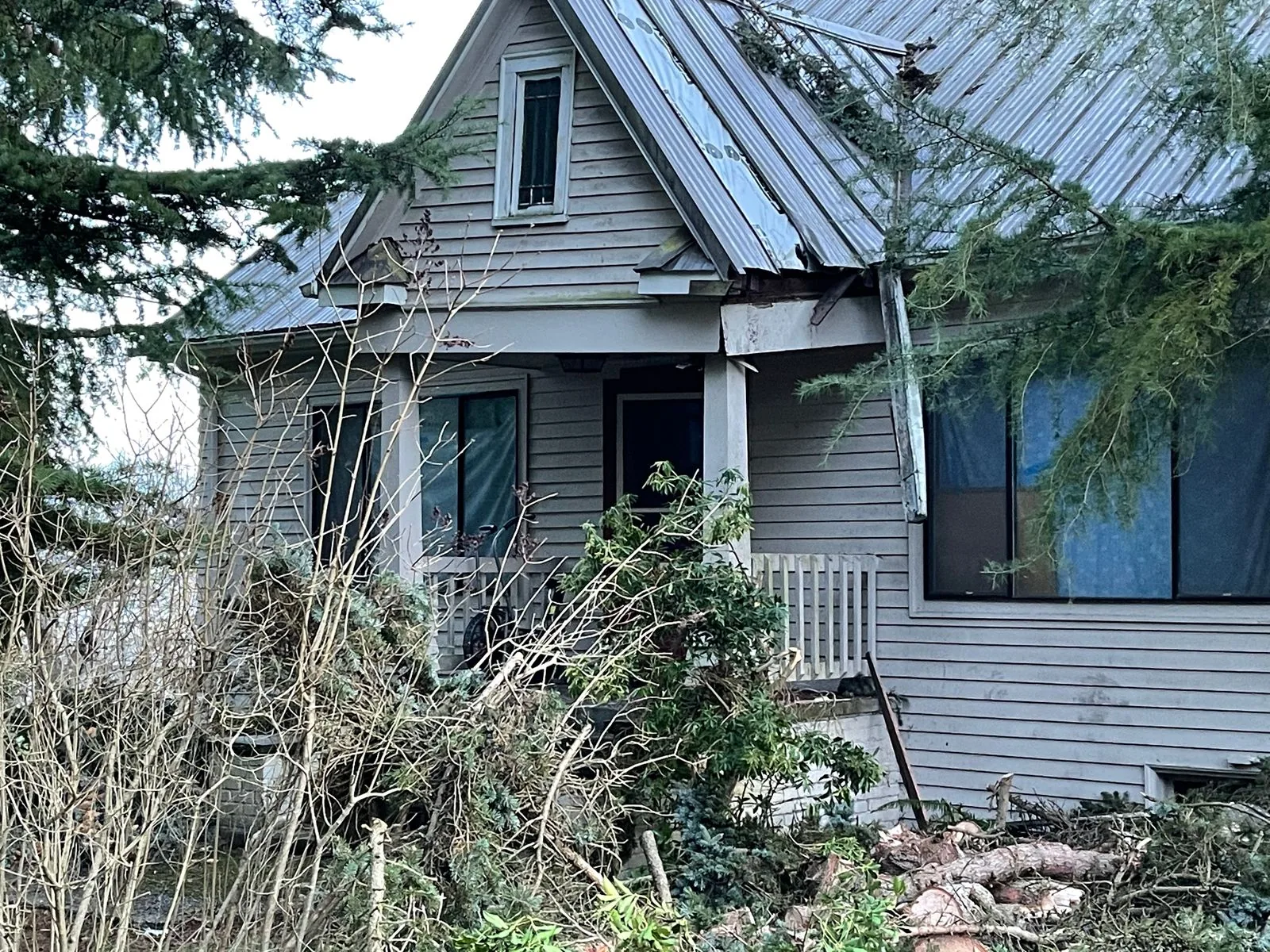 Storm-damaged older farmhouse with a broken metal roof and downed limbs in front